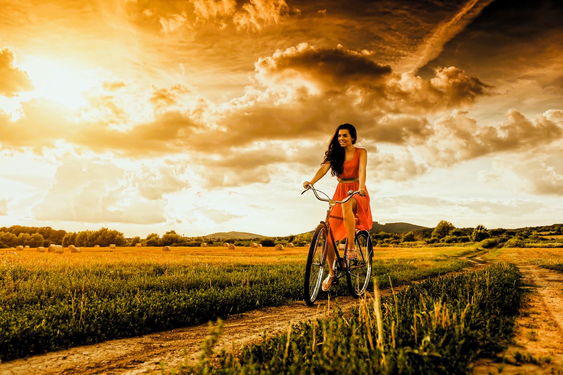A woman in a red dress rides a bicycle on a dirt path through a grassy field under a dramatic, golden sky with clouds and sun. Hills and trees are visible in the background.