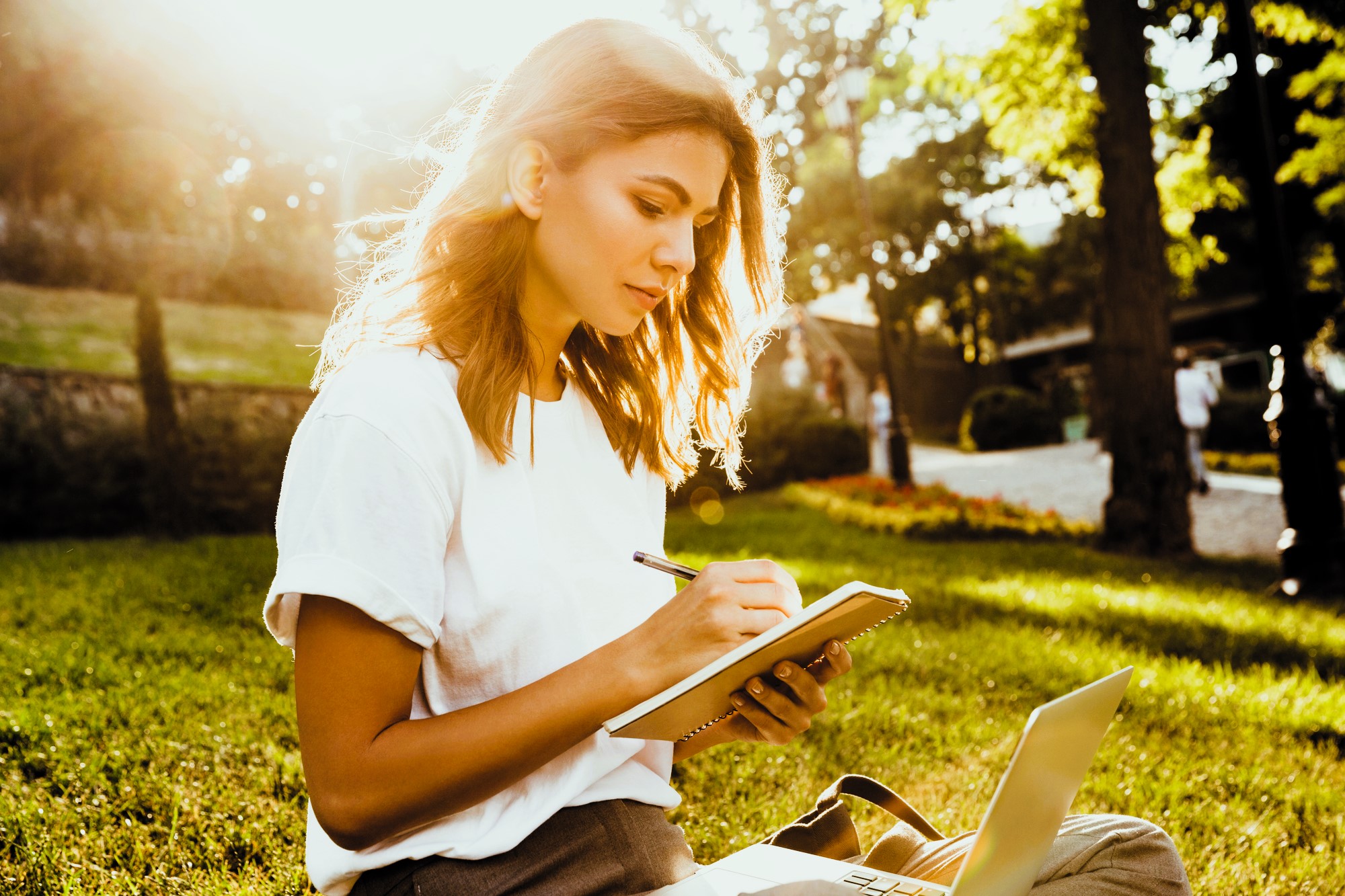 A person with long hair sits on grass in a park, writing in a notebook. They wear a white t-shirt and are next to an open laptop. Sunlight filters through trees, creating a bright, serene atmosphere.
