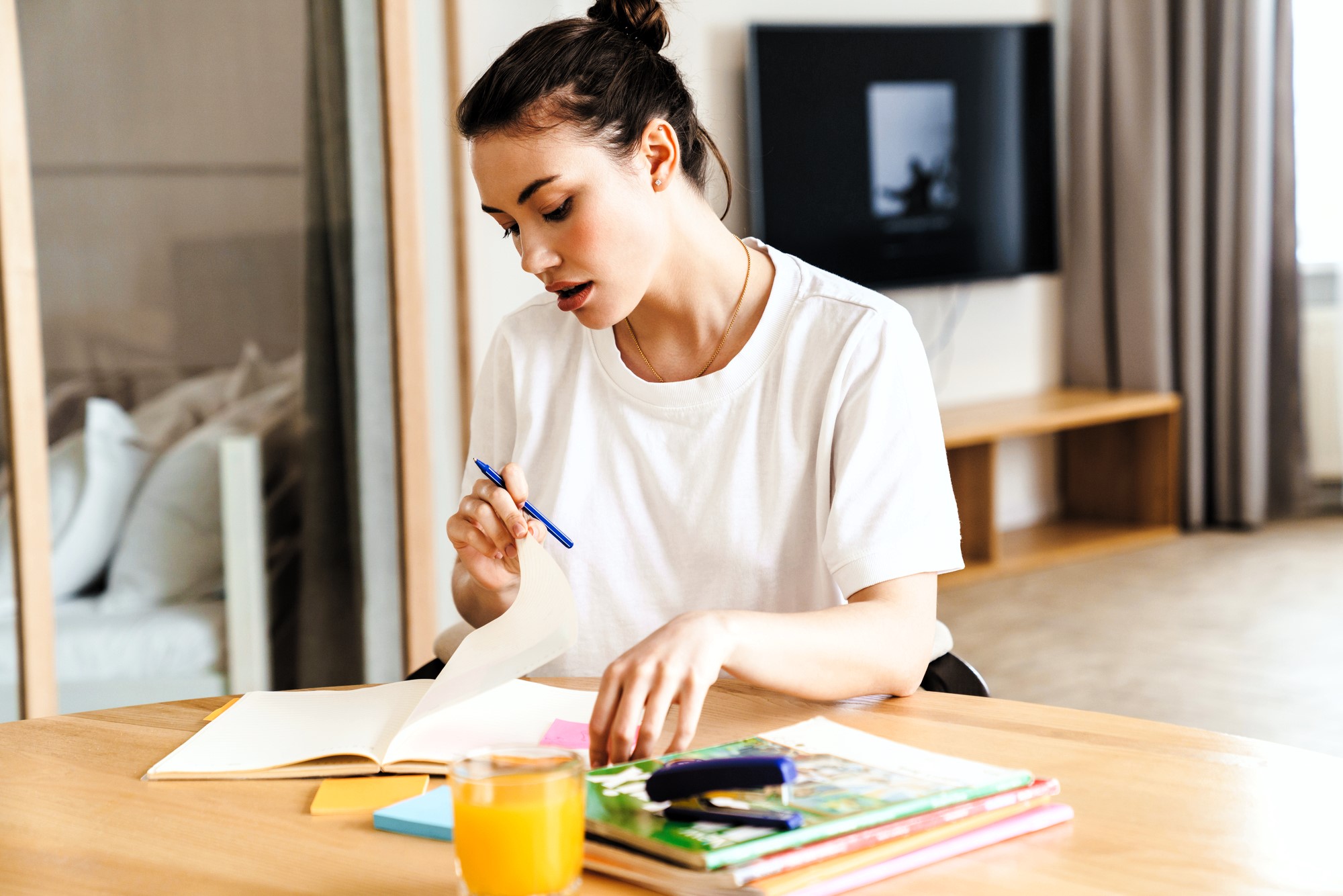 A woman with dark hair in a bun is sitting at a wooden table, writing in an open notebook with a blue pen. She has several colorful books and a glass of orange juice in front of her. A TV is visible in the background.