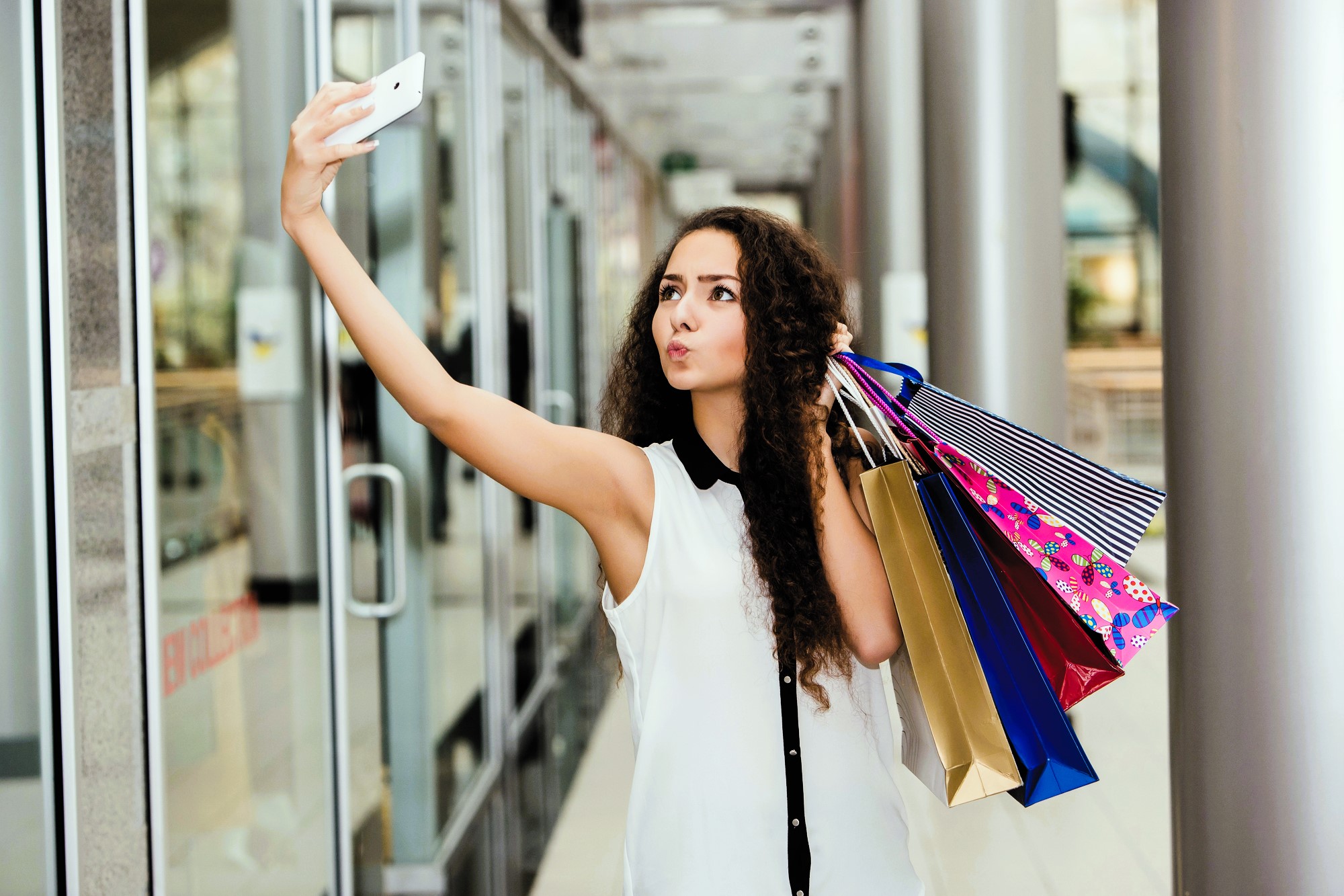 A woman with long curly hair takes a selfie while holding several colorful shopping bags in a modern mall. She wears a sleeveless blouse and stands in a corridor with glass walls and metallic columns.