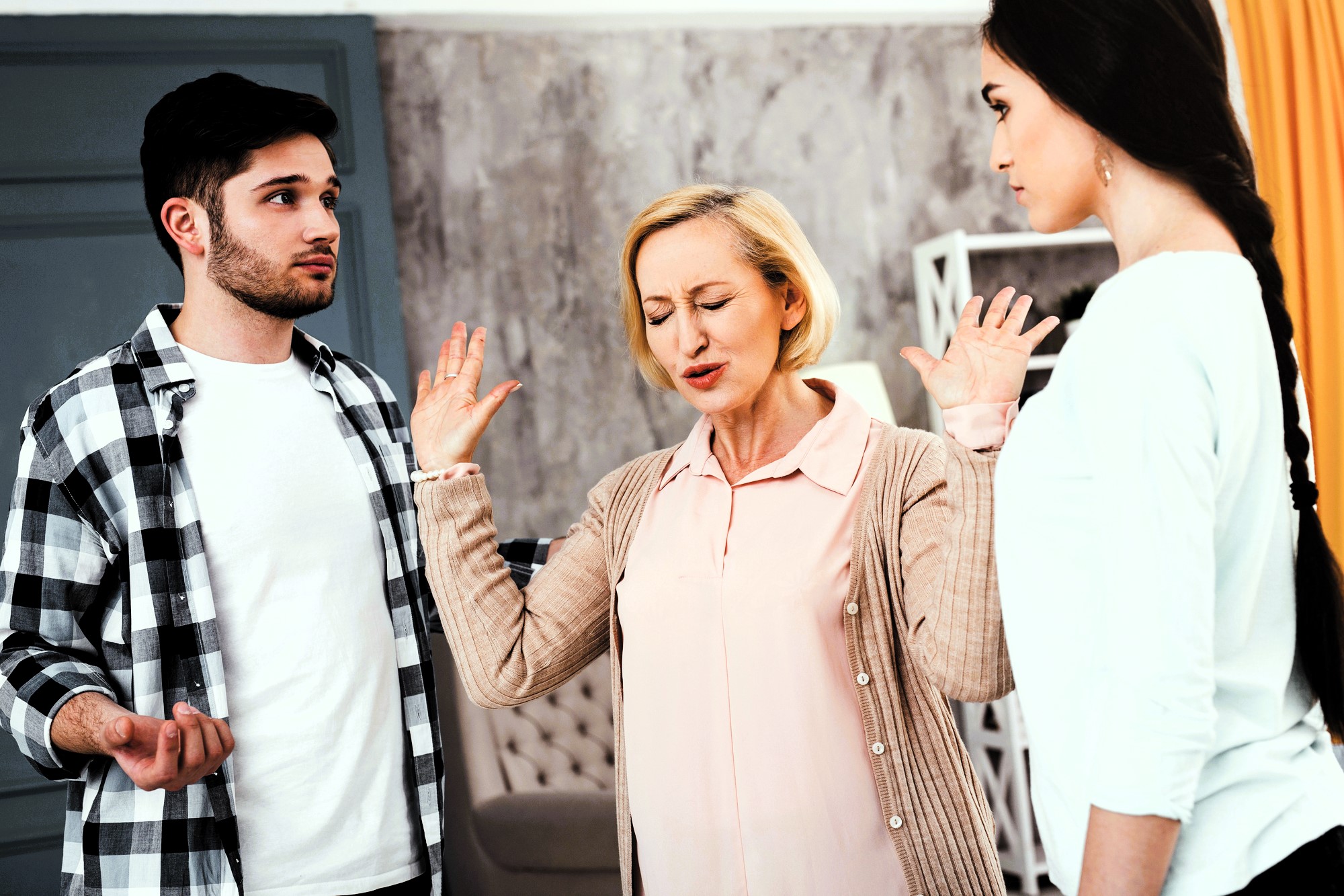 A middle-aged woman raises her hands in frustration between a young man in a plaid shirt and a young woman in a white shirt. They appear to be having a tense discussion in a room with gray walls and yellow curtains.