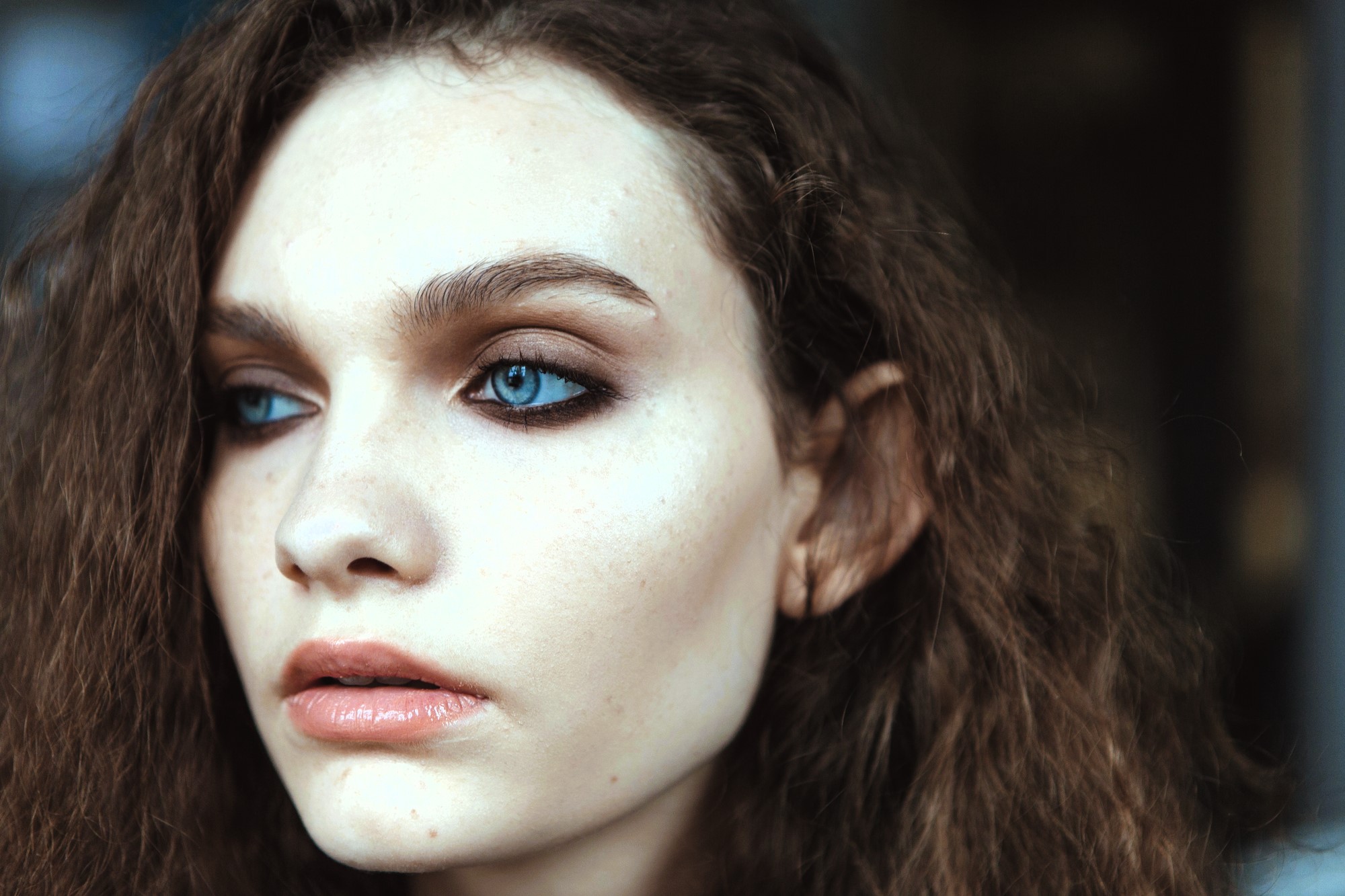 A close-up of a person with wavy brown hair and bright blue eyes, looking to the side. The lighting highlights smooth skin and subtle makeup, emphasizing the eyes and lips. The background is softly blurred.