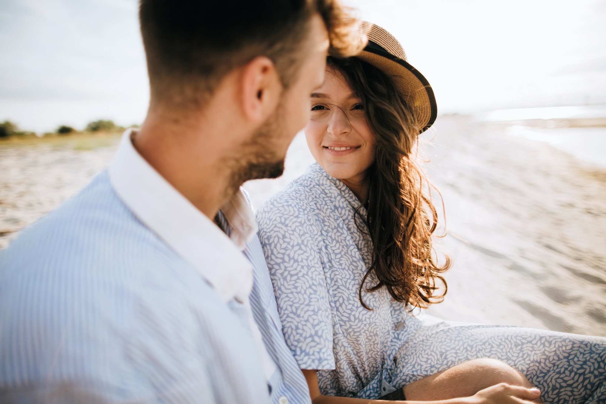 A couple sitting on a sandy beach. The woman with long brown hair is wearing a light patterned outfit and a hat, smiling at the man beside her. The man is wearing a blue and white striped shirt, and they're both enjoying a sunny day.