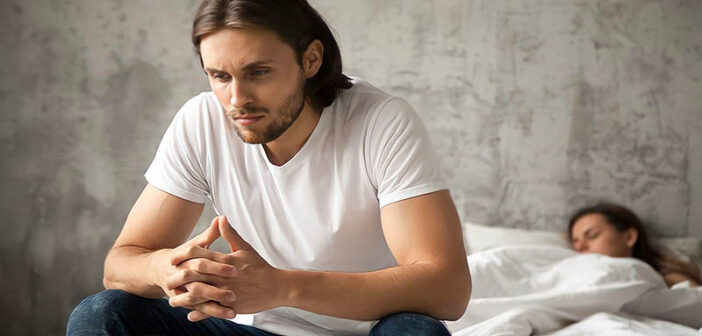 man annoyed within himself for cheating on his girlfriend with another girl who is in bed in the background