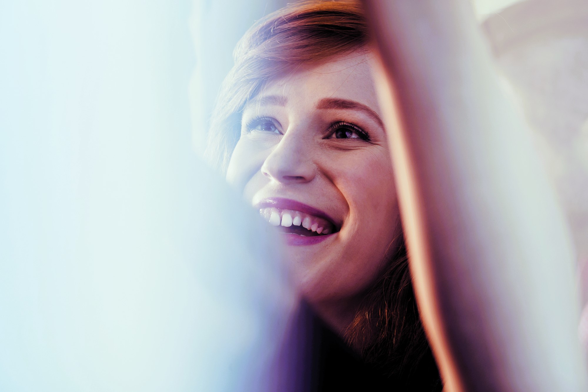A person with light brown hair is smiling widely and looking upward. The image has a soft light effect, giving a warm and cheerful ambiance. Arms are raised, partially visible in the foreground.