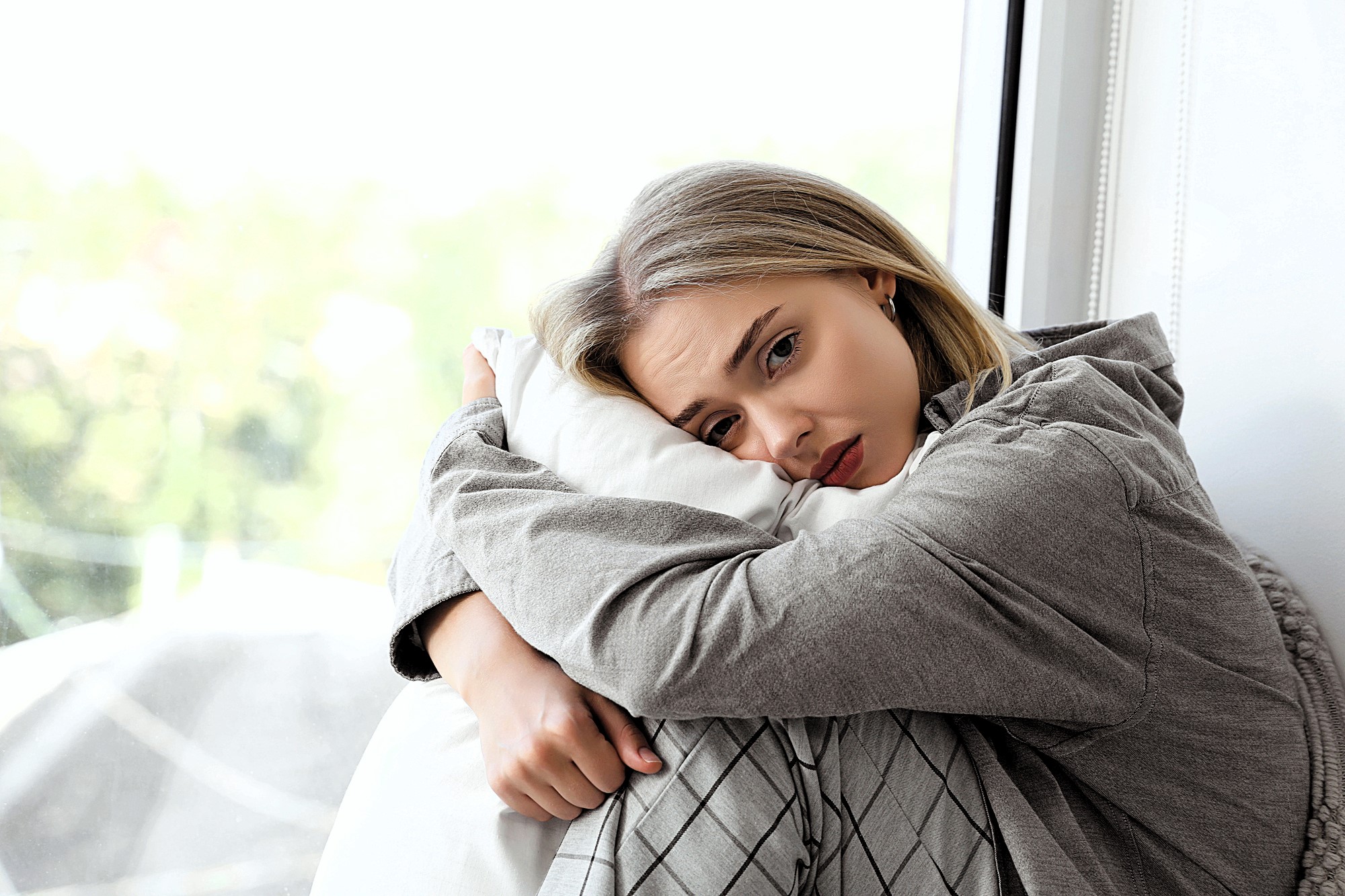 A woman with long blonde hair sits by a window, hugging a pillow. She is wearing a gray shirt and plaid pants, gazing thoughtfully outside. The background shows a blurred view of greenery, suggesting a peaceful, reflective setting.