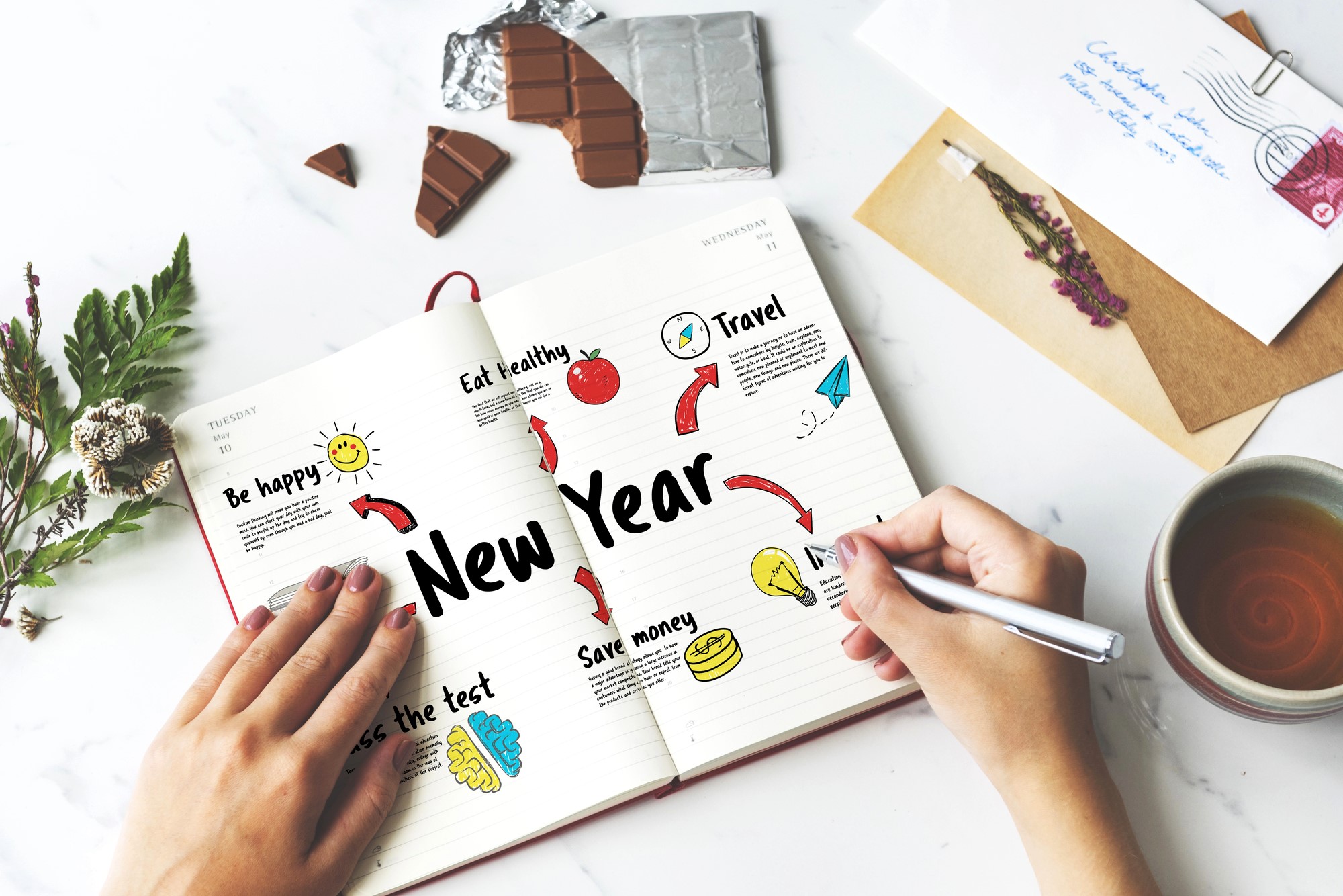 Hands holding a notebook open to a New Year goals page. Goals include "Be happy," "Eat healthy," "Travel," "Save money," and "Pass the test," accompanied by colorful sketches. Nearby are chocolate pieces, an envelope, and a cup of tea.