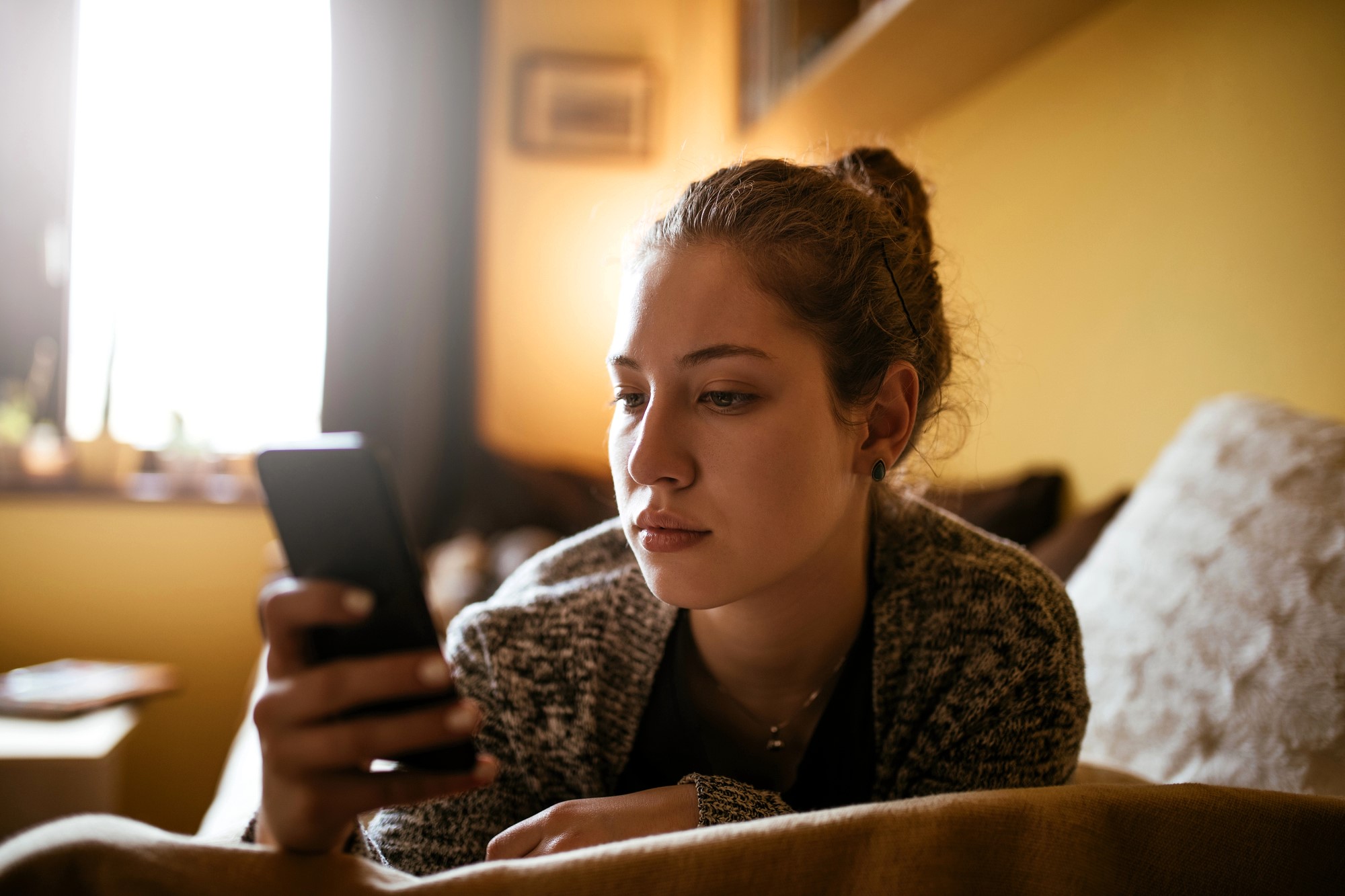 A woman with a bun hairstyle is sitting on a sofa, holding and looking at a smartphone. The background is softly lit, with a window partially visible, creating a cozy atmosphere in the room.