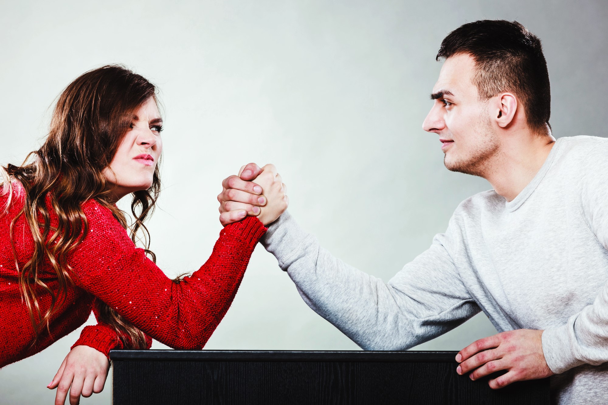 A woman in a red sweater and a man in a white sweater arm-wrestle across a table. Both are focused, with the woman showing a determined expression. The background is plain gray.