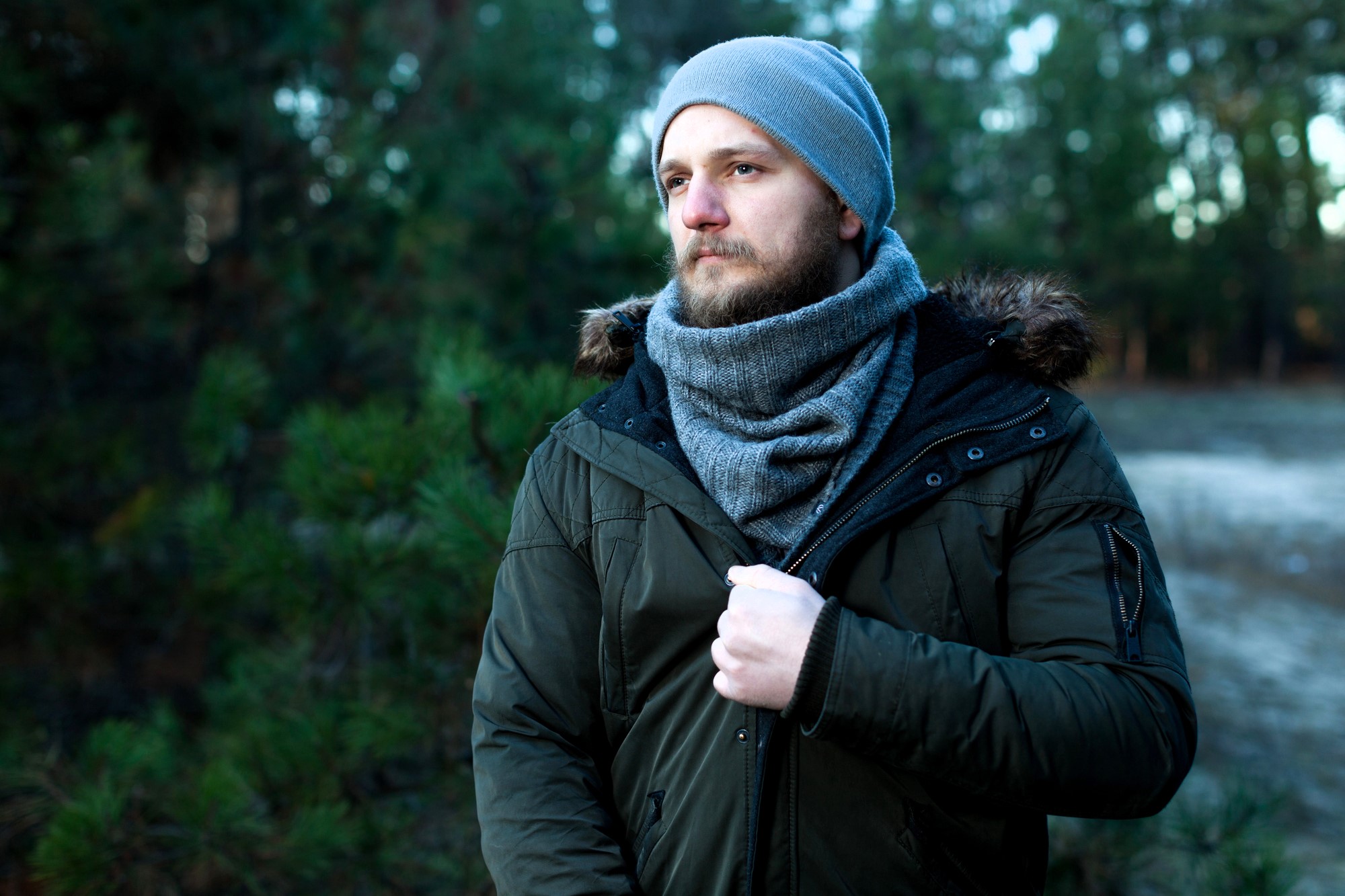 A bearded man wearing a gray beanie and scarf stands outside in a forested area. He is dressed in a green winter jacket with faux fur trim. He gazes thoughtfully into the distance, with trees blurred in the background.