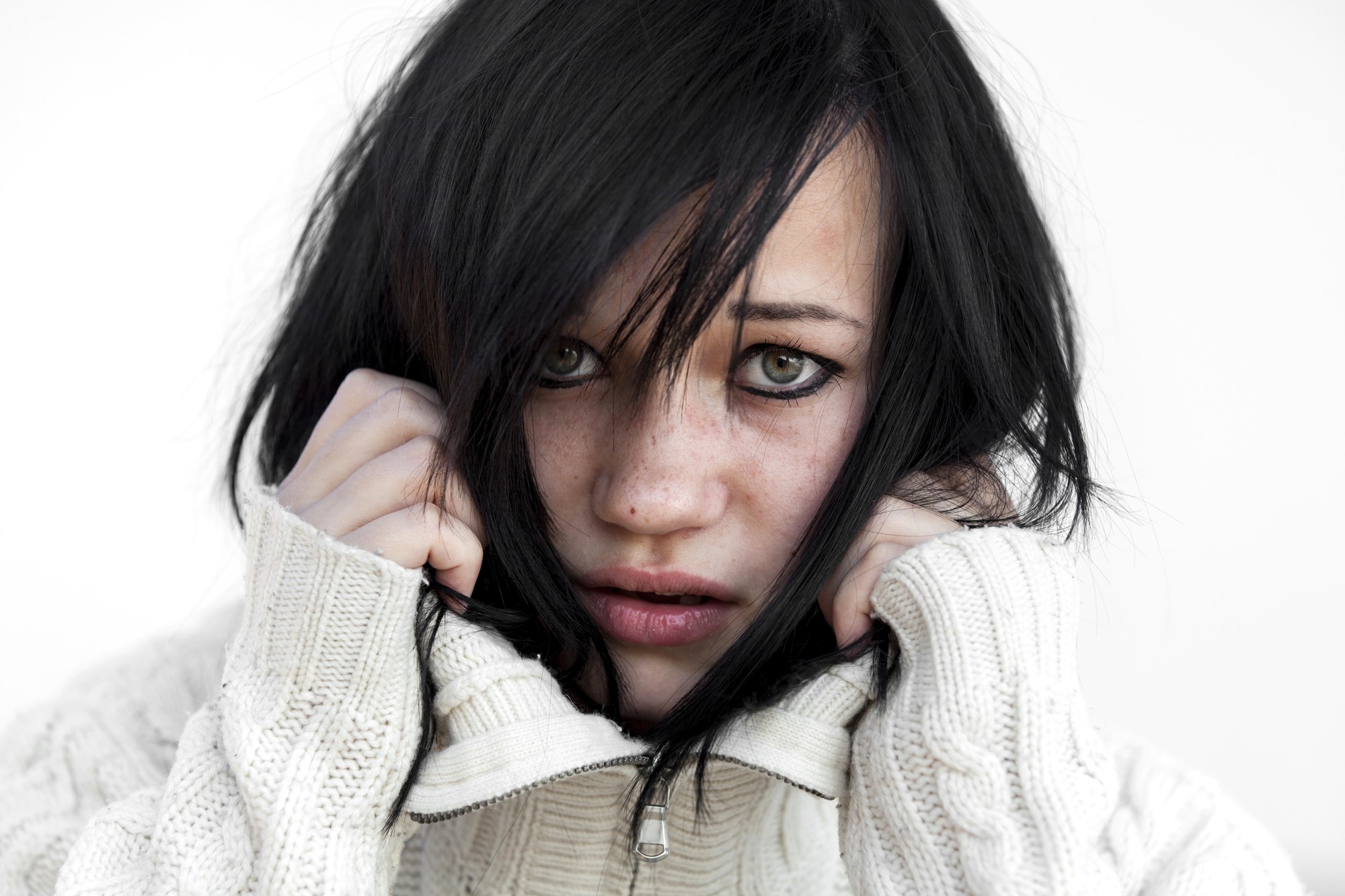 A person with black hair, wearing a white cable-knit sweater, covers part of their face with the sweater's collar. They have an intense expression, with hair partially falling over their eyes, against a plain white background.