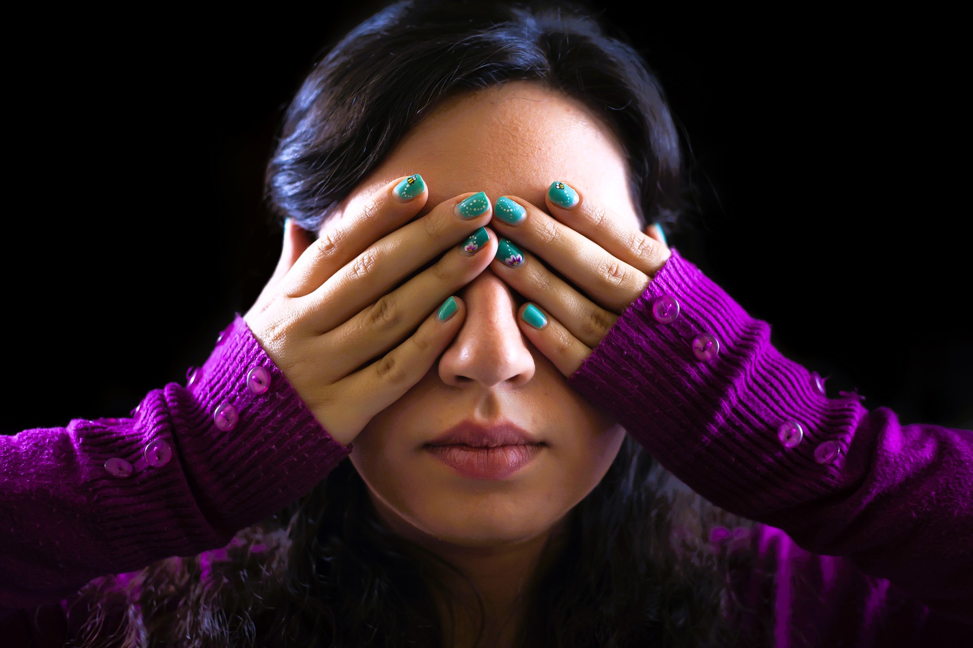 A woman with long dark hair covers her eyes with her hands. She has turquoise nail polish and wears a purple sweater with button details on the sleeves. The background is black.