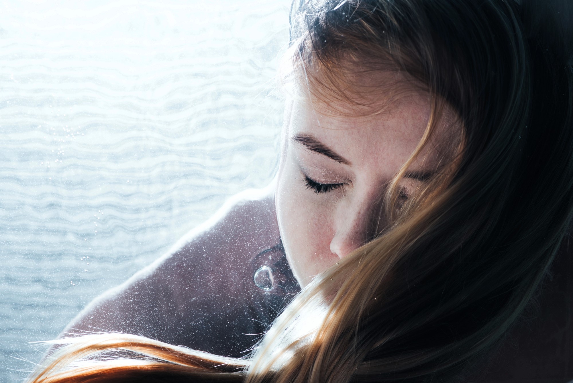 A person with long hair appears submerged in water, eyes closed, and surrounded by light bubbles. Sunlight filters through, casting a serene and ethereal glow across their peaceful expression.
