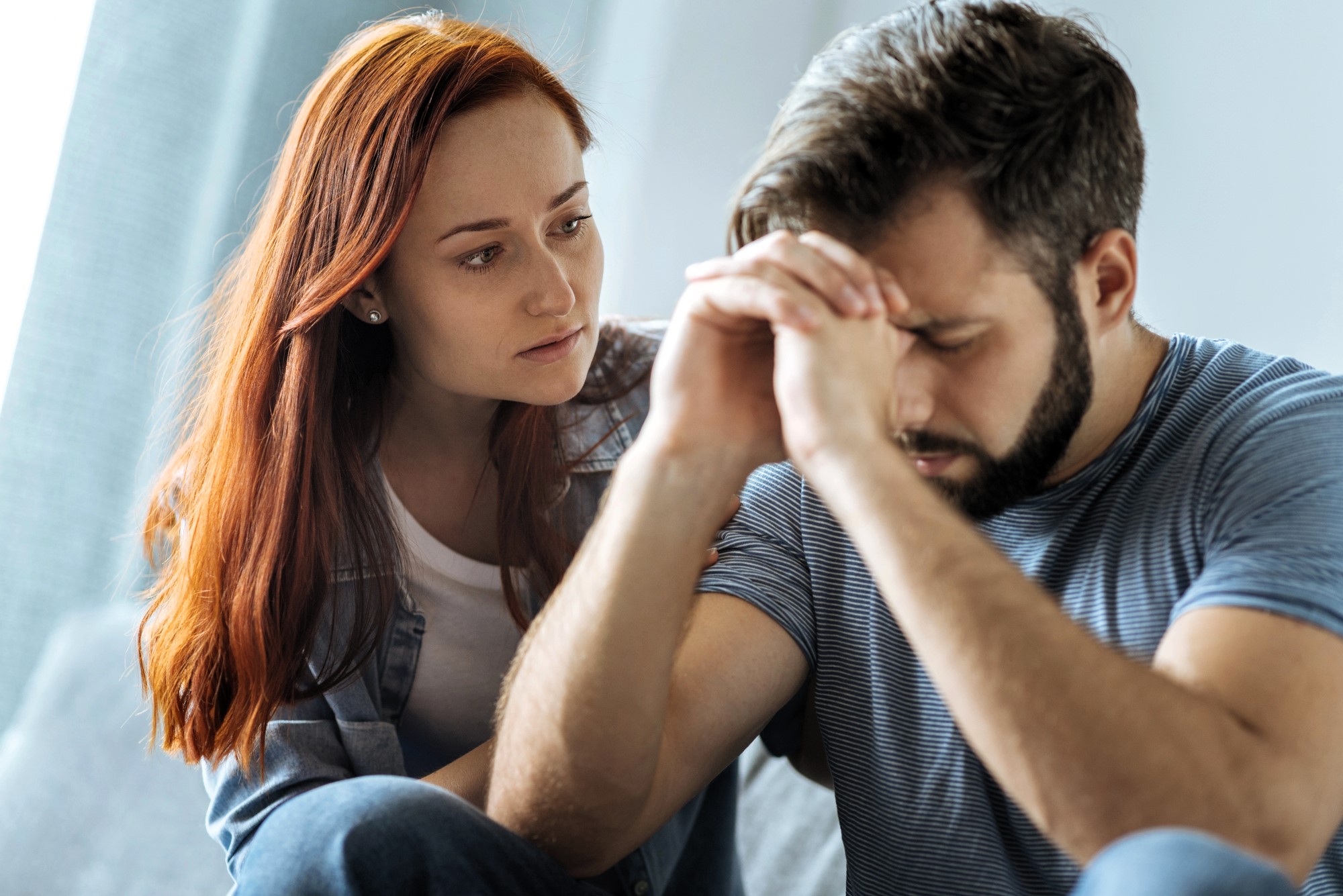 A woman with red hair looks concerned and supportive as she sits next to a man with brown hair, who appears upset and is resting his head on his hands. They are indoors, and he seems to be experiencing distress.