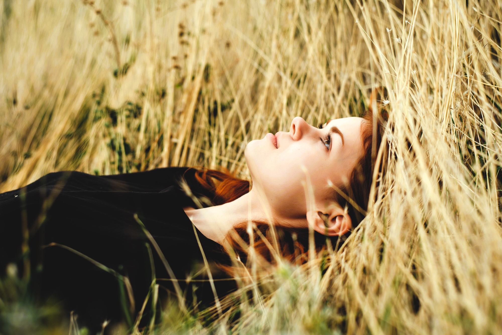 A person with long hair lies in a field of tall, dry grass, gazing upward. They are wearing a dark shirt, and the scene conveys a sense of tranquility and contemplation amidst nature.
