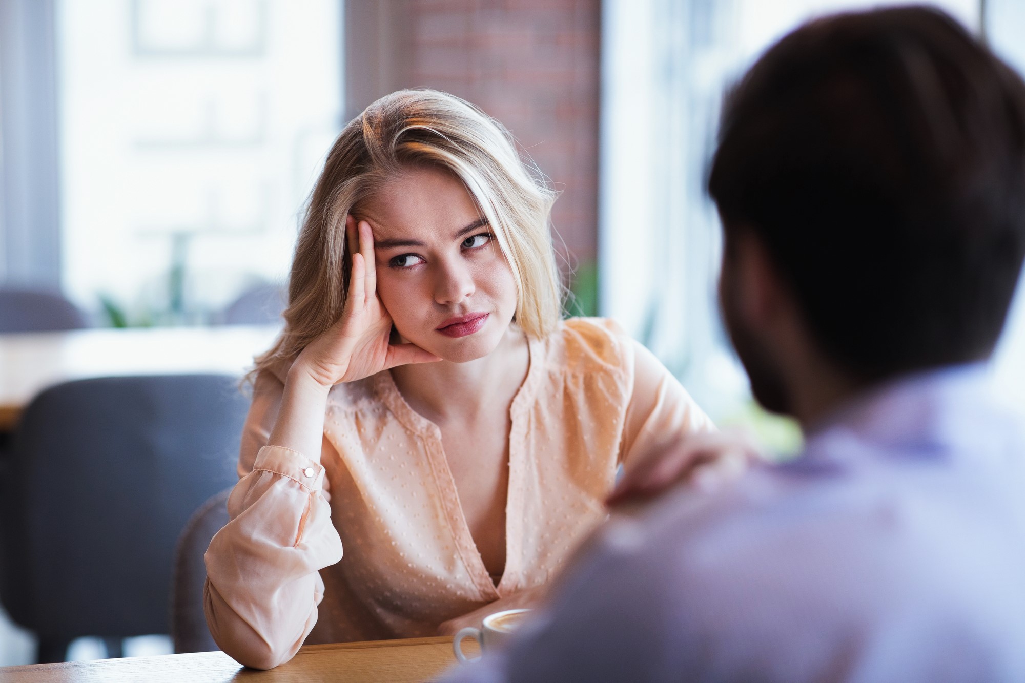 A woman with blonde hair is sitting at a table, resting her head on her hand, looking bored or frustrated. She is wearing a light peach blouse and is facing a man whose back is to the camera. They're in a bright indoor setting.