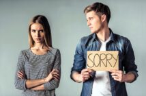 A man in a denim shirt holds a cardboard sign that says "SORRY," looking at a woman in a striped shirt who appears upset with crossed arms. They stand against a plain gray background.