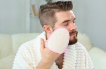 man in fluffy bath robe using exfoliating glove on his face - illustrating a feminine husband