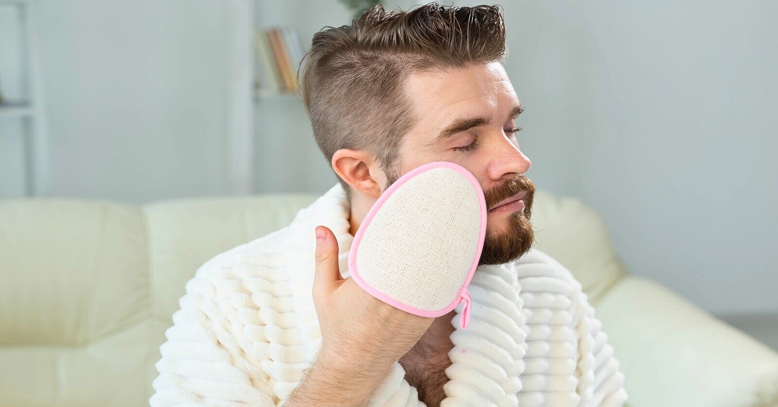 man in fluffy bath robe using exfoliating glove on his face - illustrating a feminine husband