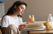 young woman sips orange juice while reading a book at the coffee table