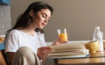 young woman sips orange juice while reading a book at the coffee table