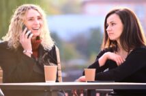 two friends sat at an outdoor table with hot drinks - the one on the left is speaking on the phone while the one on the right has an annoyed expression on her face