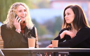 two friends sat at an outdoor table with hot drinks - the one on the left is speaking on the phone while the one on the right has an annoyed expression on her face