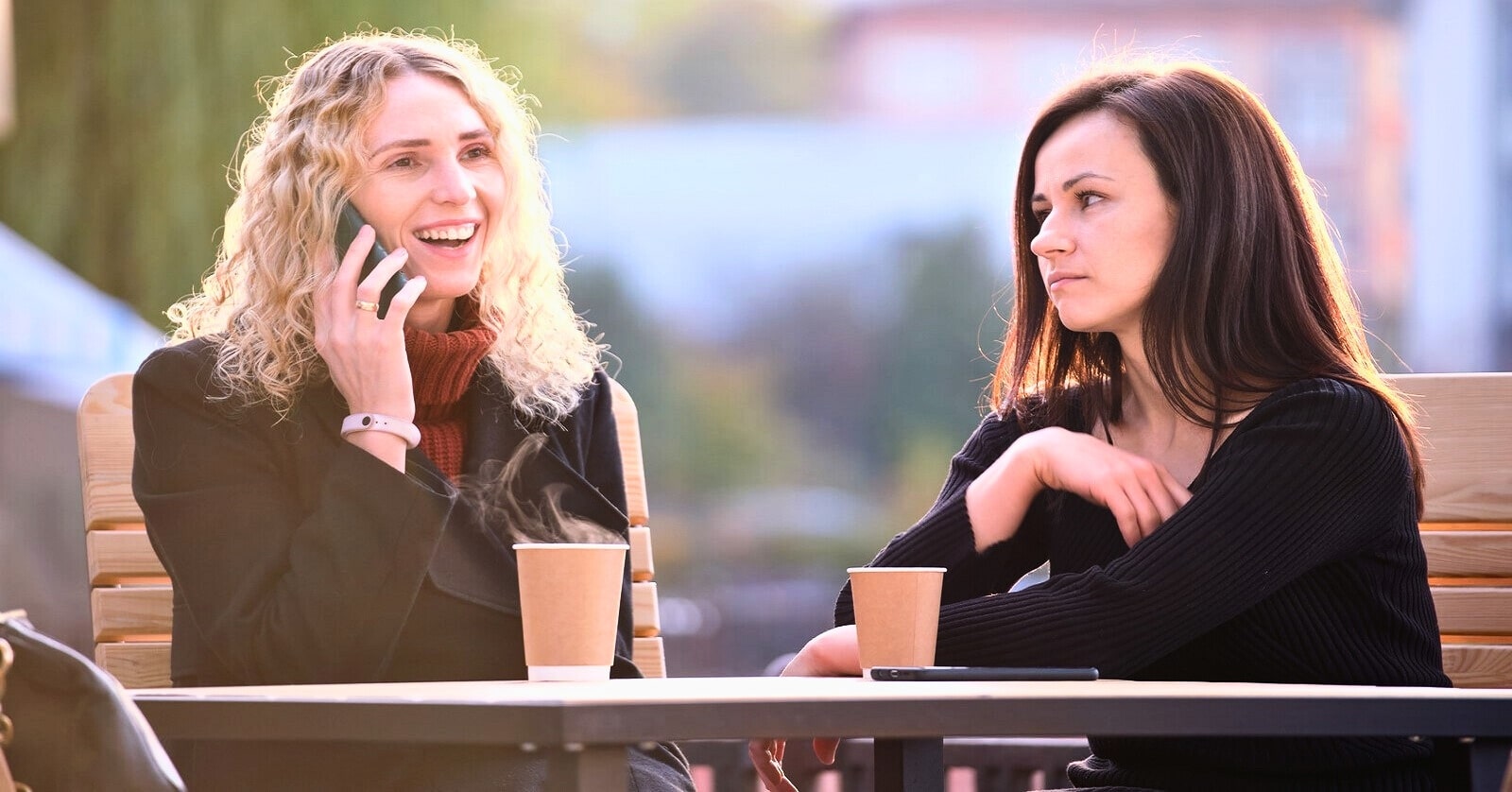 two friends sat at an outdoor table with hot drinks - the one on the left is speaking on the phone while the one on the right has an annoyed expression on her face