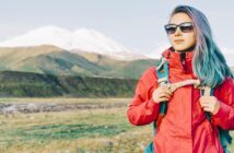 young woman with colorful hair and bright red jacket hiking across plains with a mountain in the background