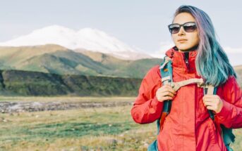young woman with colorful hair and bright red jacket hiking across plains with a mountain in the background