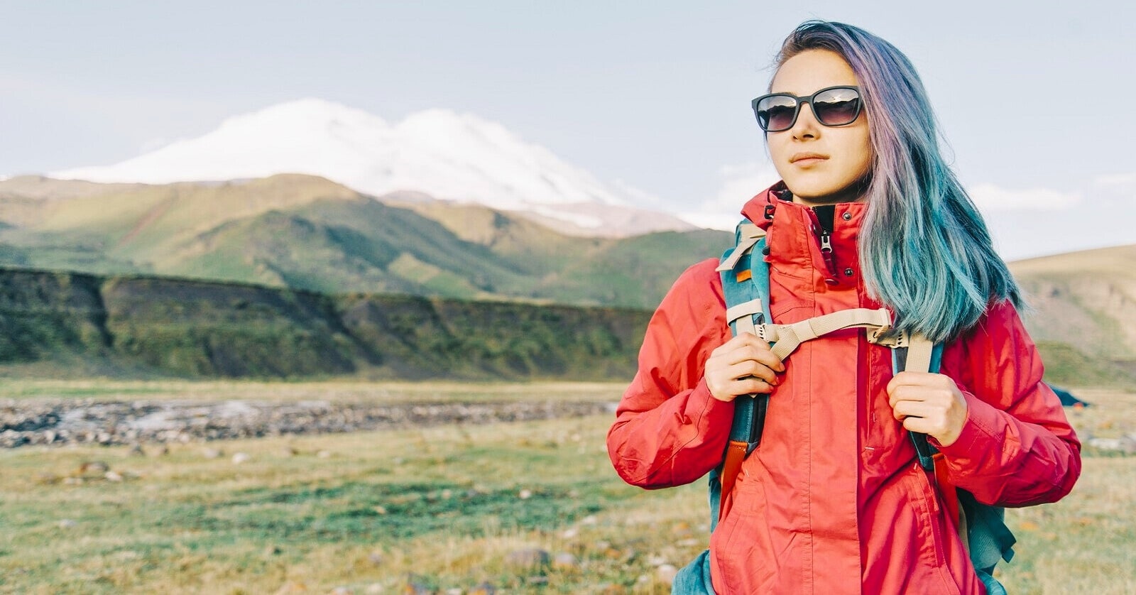 young woman with colorful hair and bright red jacket hiking across plains with a mountain in the background