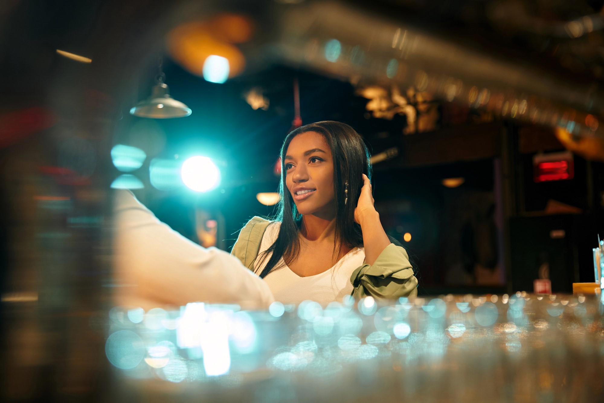 Woman with long hair sits at a bar, smiling while looking to the side. The background is softly blurred with colorful lights, creating a warm, inviting atmosphere. Glasses are visible in the foreground.