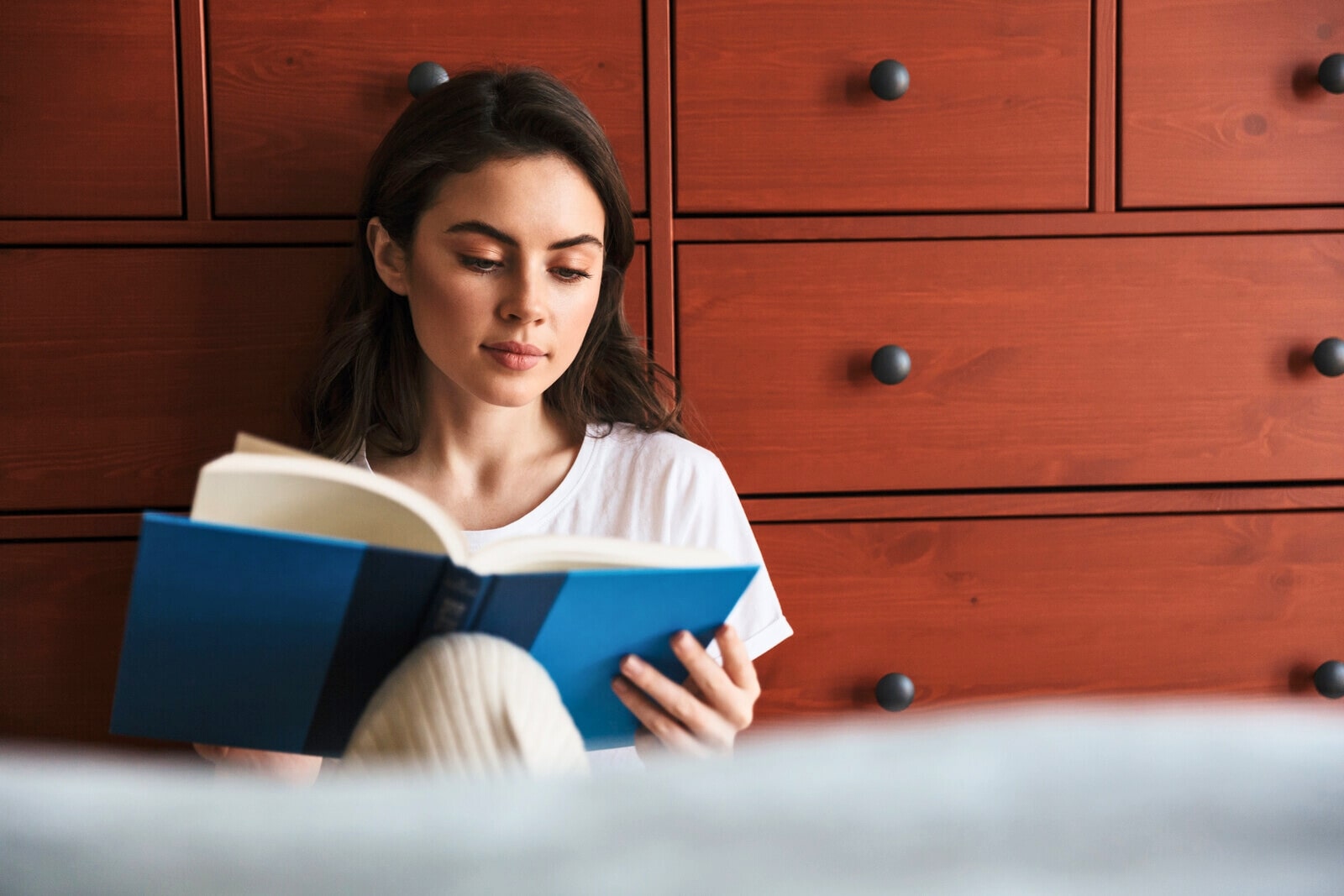 introverted woman sitting on the floor against a chest of drawers reading a book