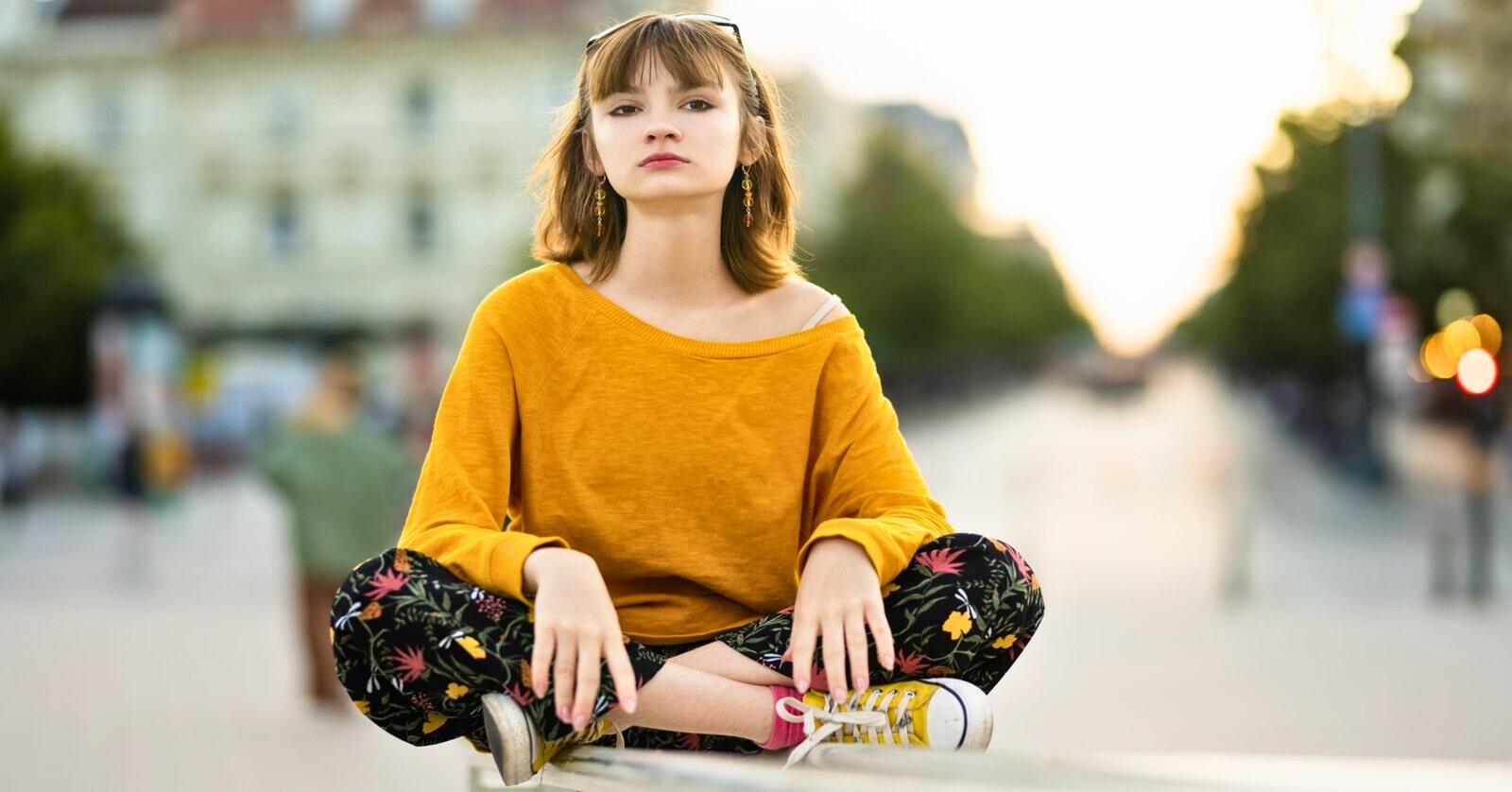 a young woman wearing a bright orange top sitting cross-legged on some handrails with a urban backdrop