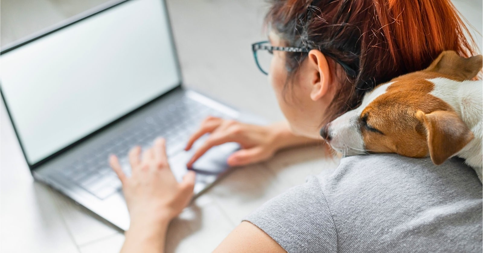 young woman with red hair laying on floor working on her laptop while her small dog sits on her back