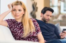 woman looking discontent as her partner looks at his phone on the couch behind her - illustrating incompatibility in a relationship
