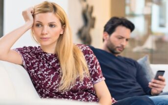 woman looking discontent as her partner looks at his phone on the couch behind her - illustrating incompatibility in a relationship