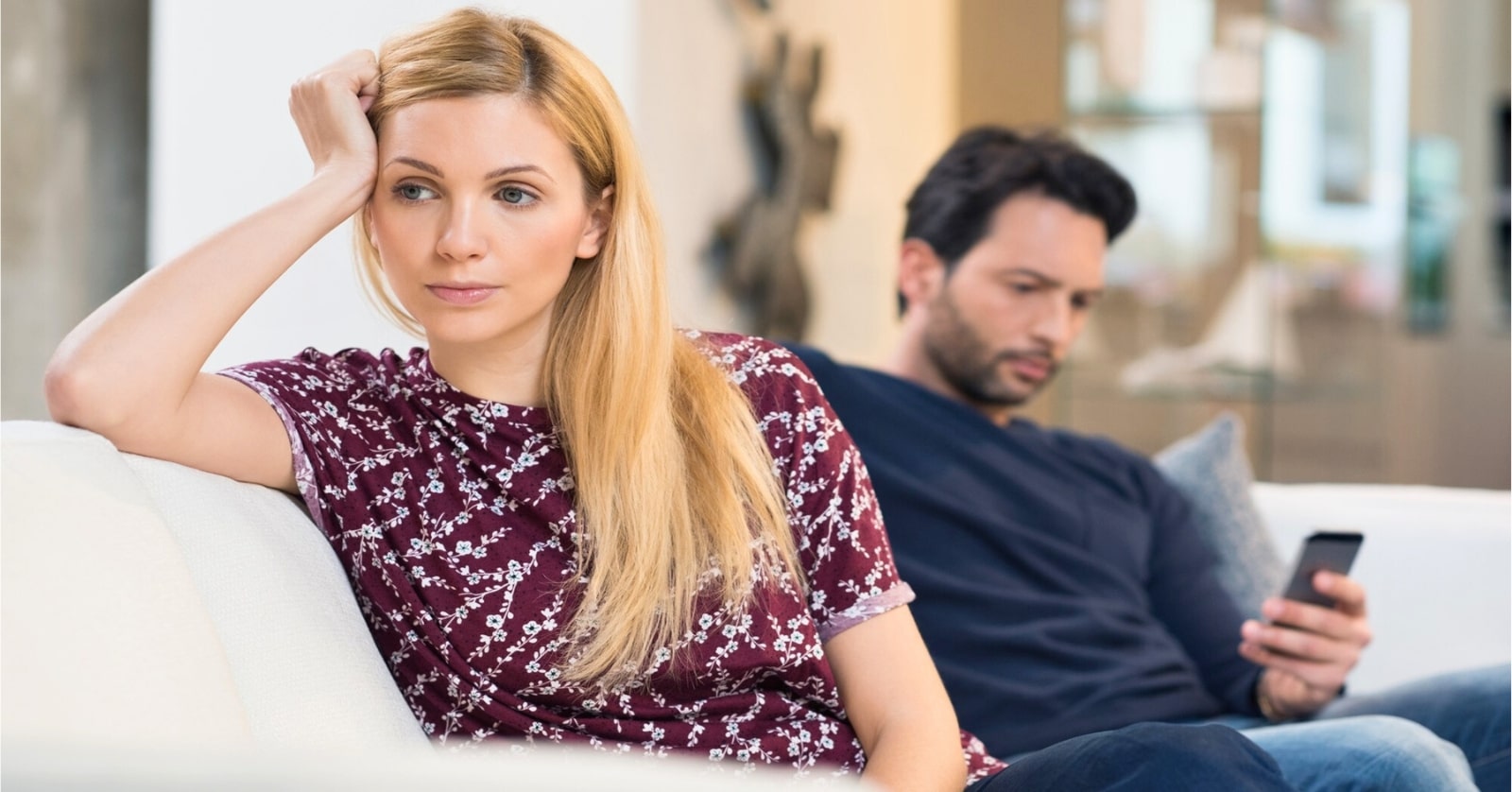 woman looking discontent as her partner looks at his phone on the couch behind her - illustrating incompatibility in a relationship