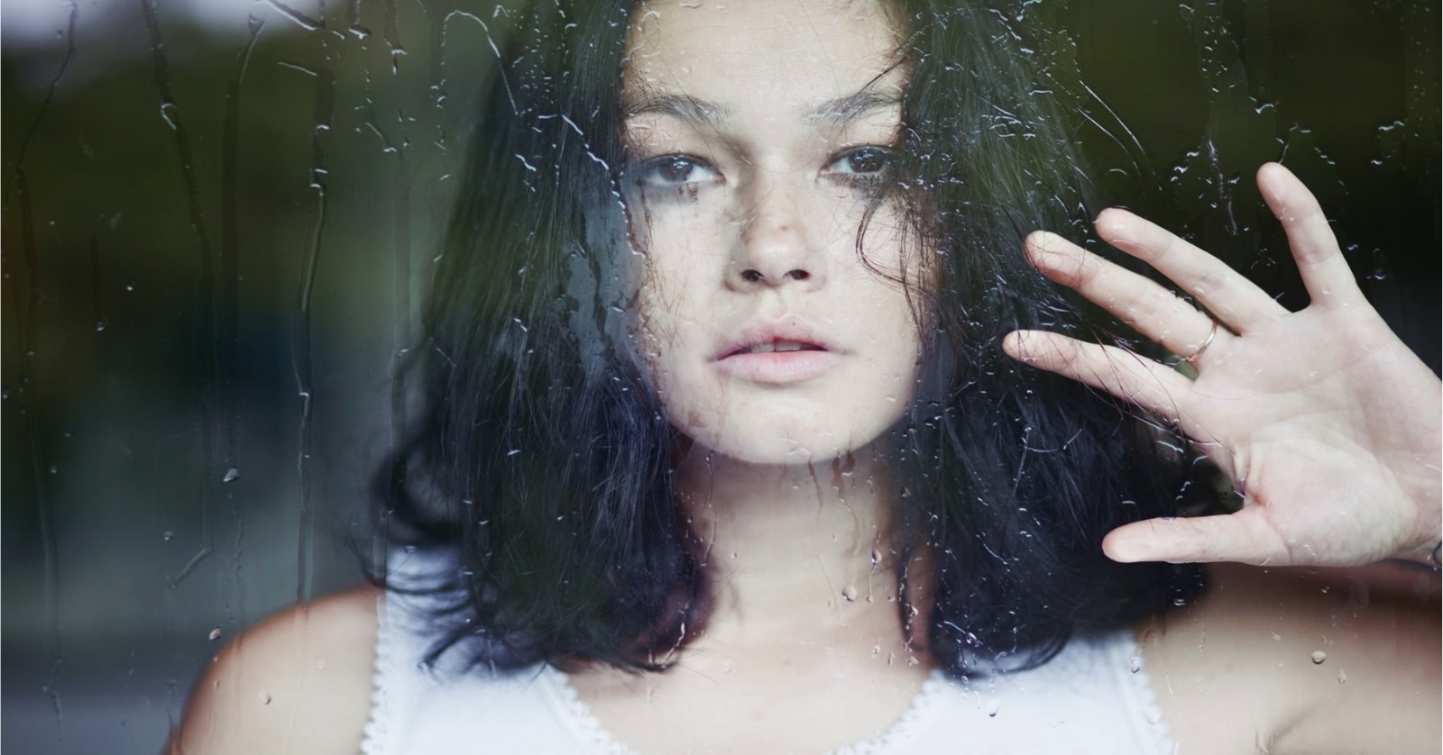 pensive looking young woman facing camera through a glass window - she has her hand pressed up against the window