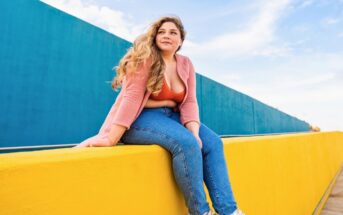 plus sized woman sitting on a bright yellow wall against a blue backdrop, looking and feeling confident in her body