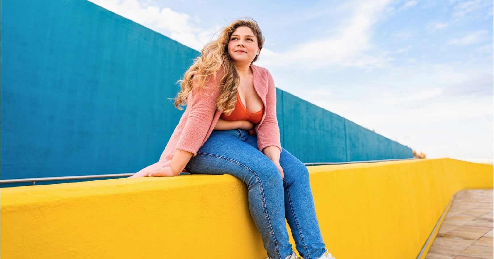 plus sized woman sitting on a bright yellow wall against a blue backdrop, looking and feeling confident in her body