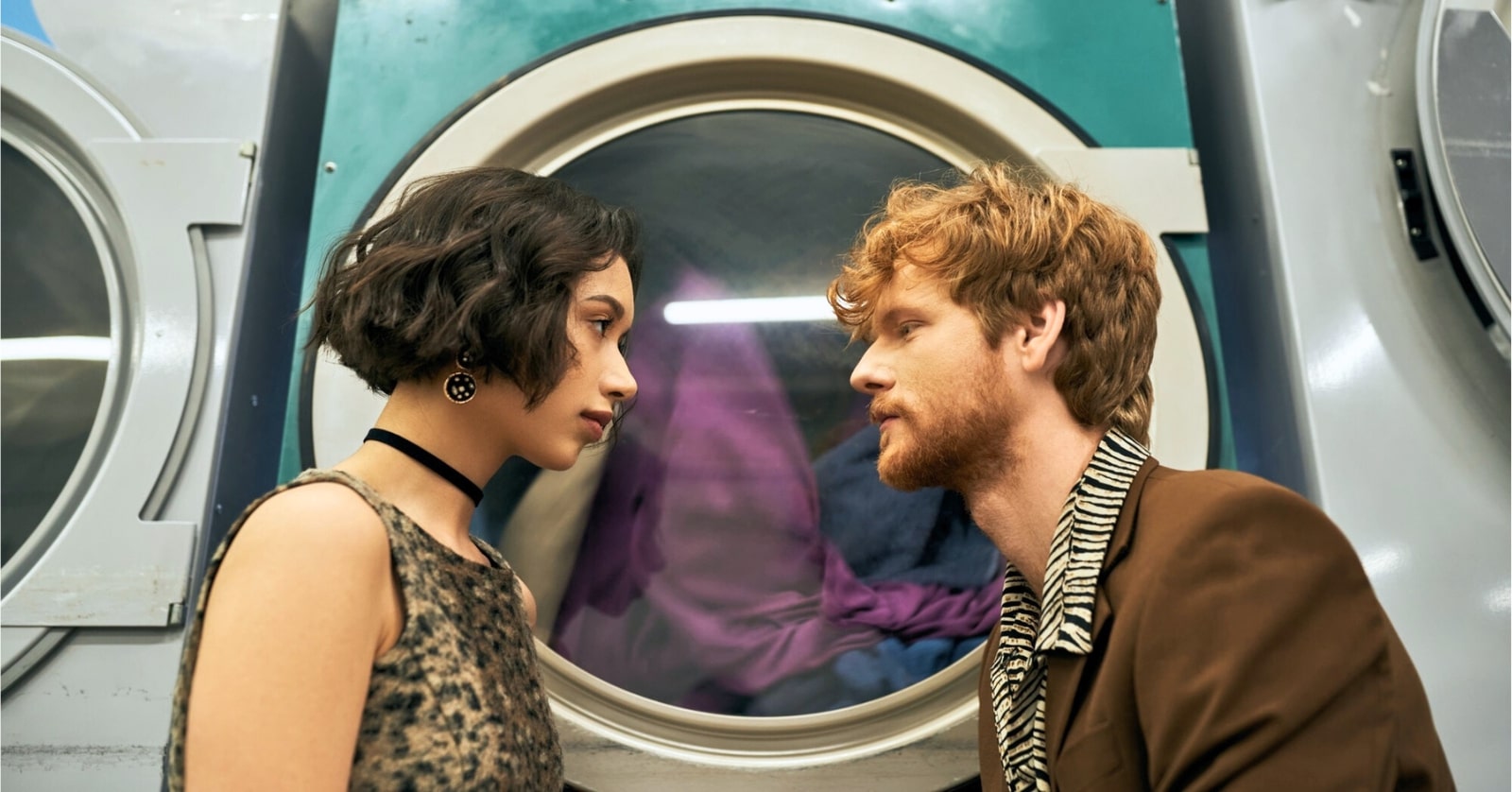 young man and young woman looking into each other's eyes in a laundromat setting with a large washing machine in the background