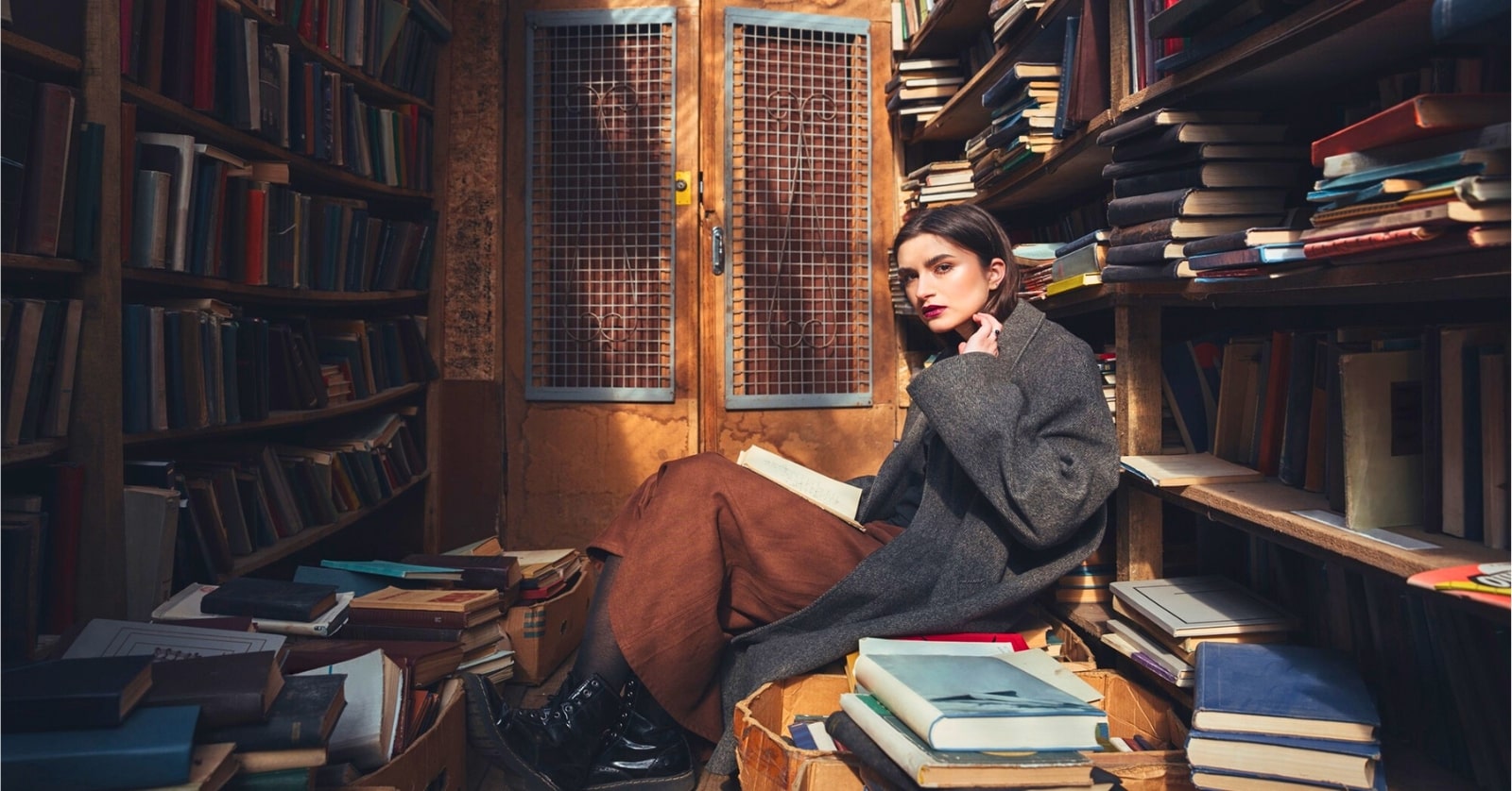 young woman sitting among hundreds of books on bookcases and piled in boxes