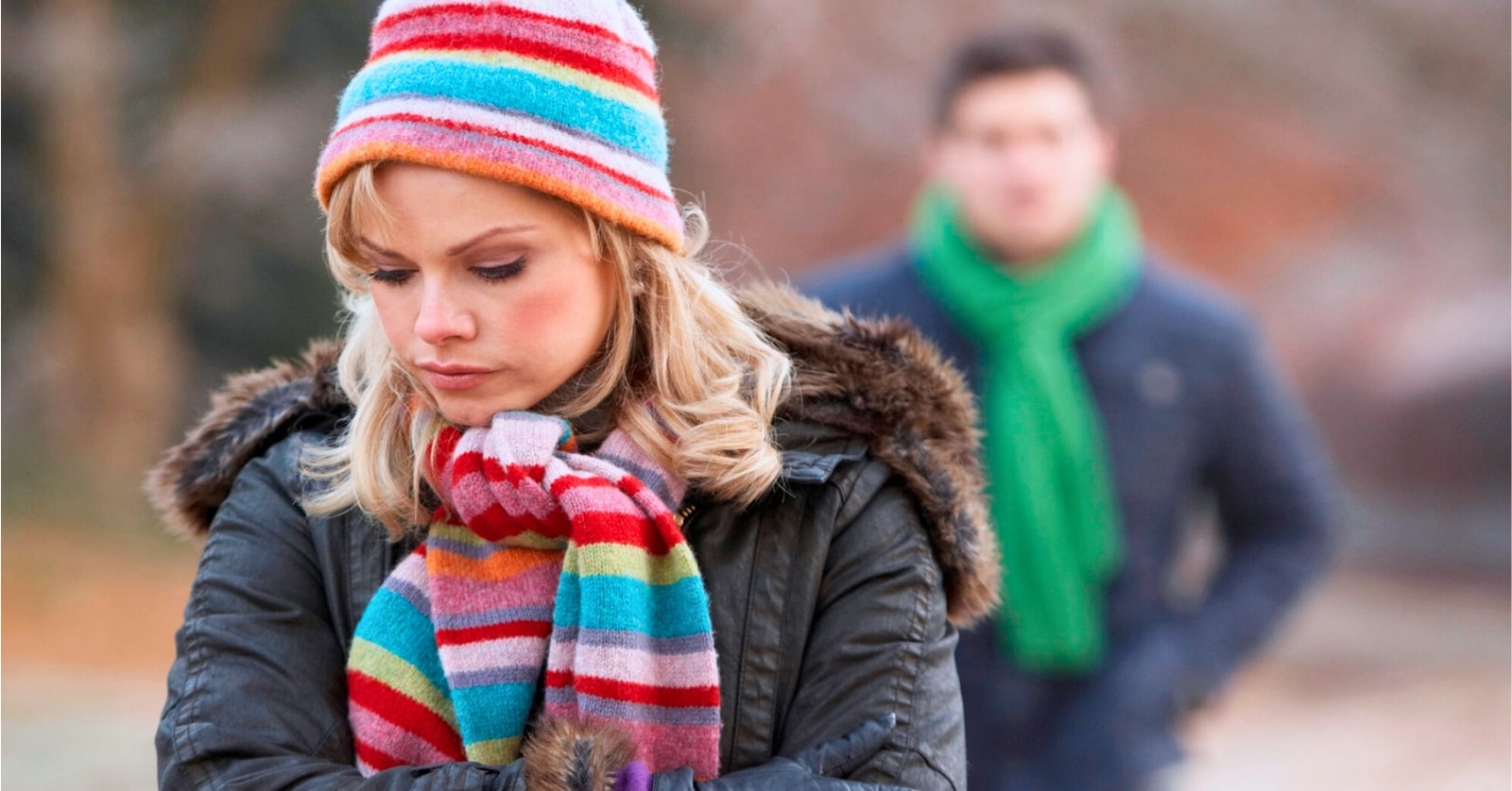 woman in the foreground wearing colorful hat and scarf with a pensive, slightly unhappy expression on her face as her boyfriend walks behind her in the distance