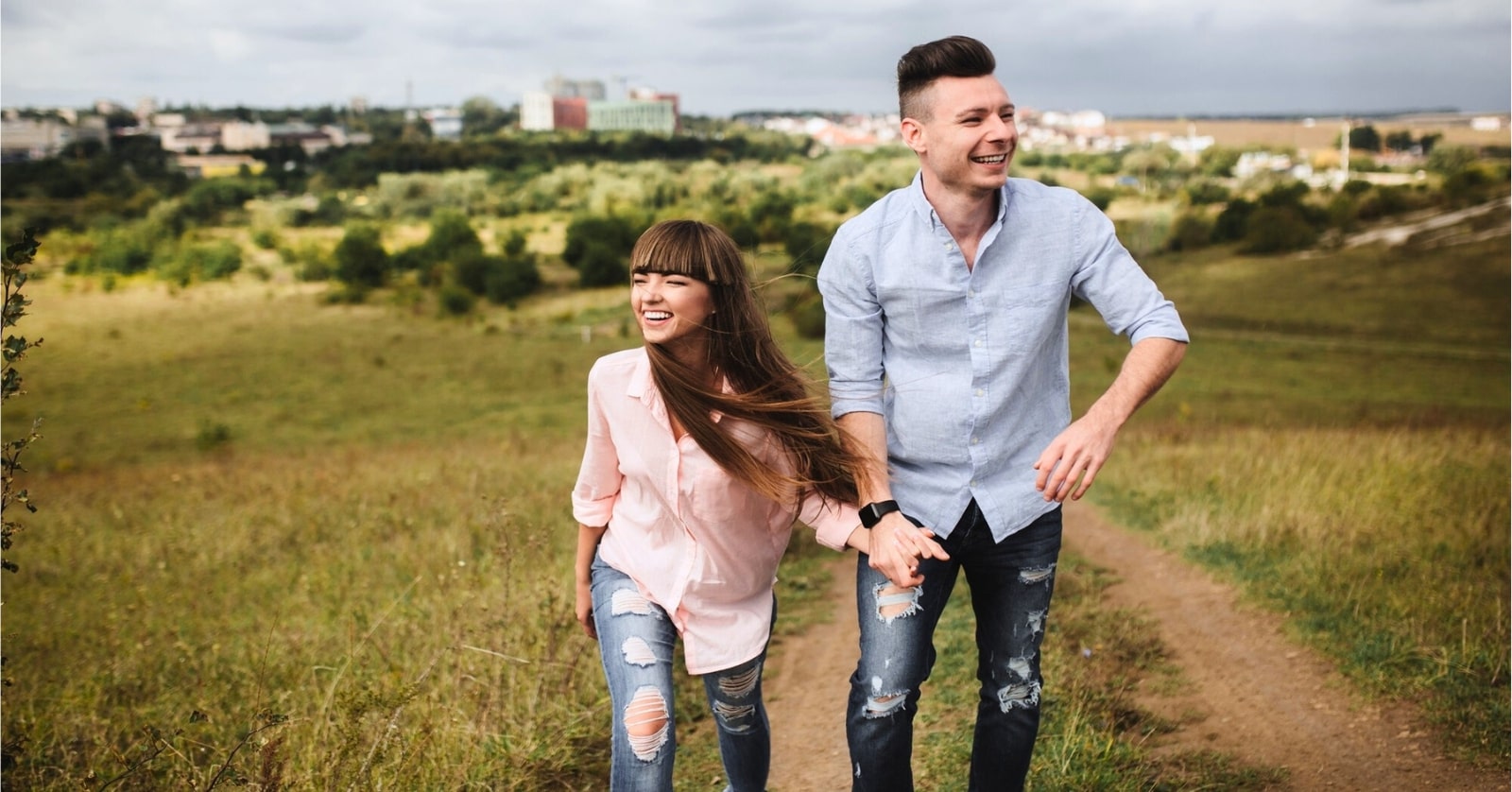 happy couple walking up a grassy hill path with a city in the background