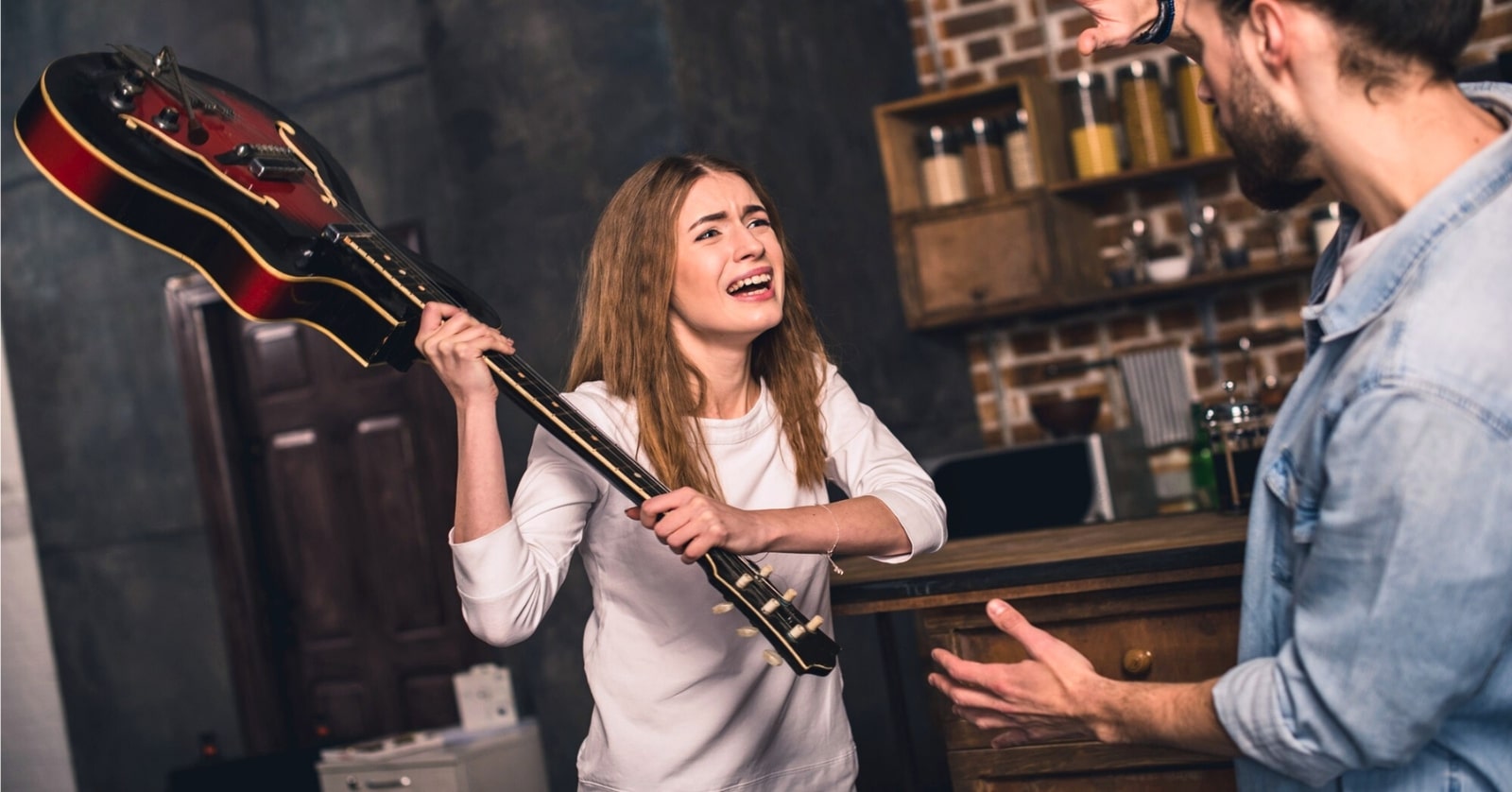 young woman with upset angry expression holding a guitar up to threaten a man with