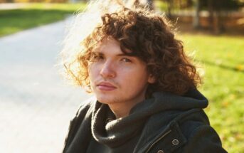 young man with curly hair sitting on a bench in a park, a pensive and forlorn expression on his face