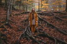 woman dressed warmly standing in the forest surrounded by large tree roots and autumnal leaves