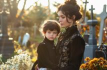 young woman holding her son, they are both dressed in black funeral attire, they are standing in a graveyard