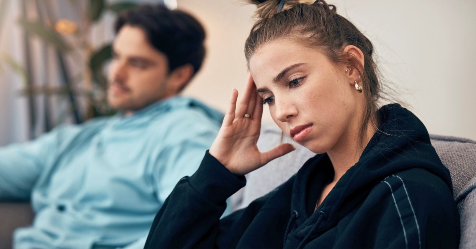 woman sitting on the couch with her boyfriend nearby, she leans her head on her hand as if to question whether she is happy in the relationship